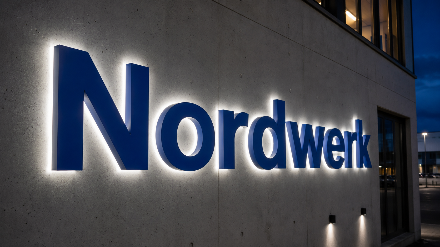 Blau leuchtendes Firmenlogo "Nordwerk" als 3D-Buchstaben an einer Betonfassade bei Dämmerung, Hintergrundfenster und Parkplatz.