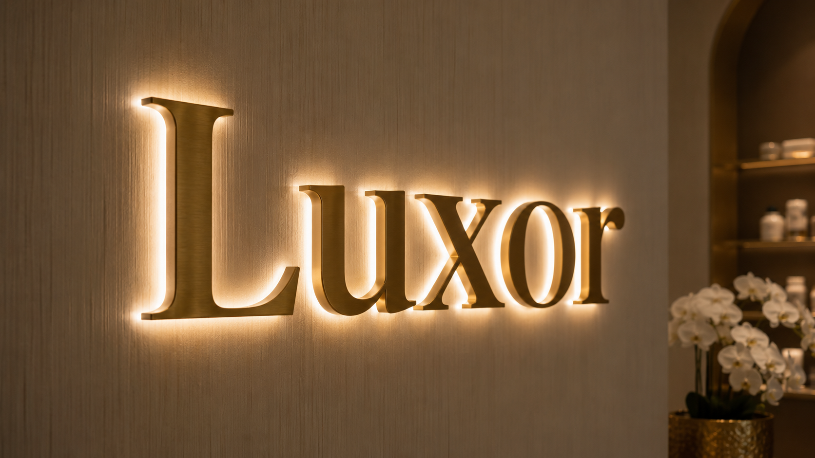 Goldene, beleuchtete Schriftzüge an einer Wand, die das Wort "Luxor" bilden, rechts Orchideen in einem Topf sichtbar.