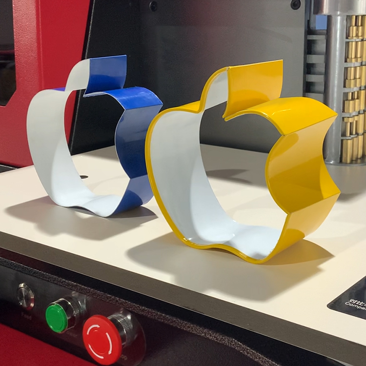 Zwei glänzende, dreidimensionale Apple-Logos aus gebogenem Metall oder Kunststoff, eines gelb und eines blau, auf einer Arbeitsplatte vor Maschinengehäuse.