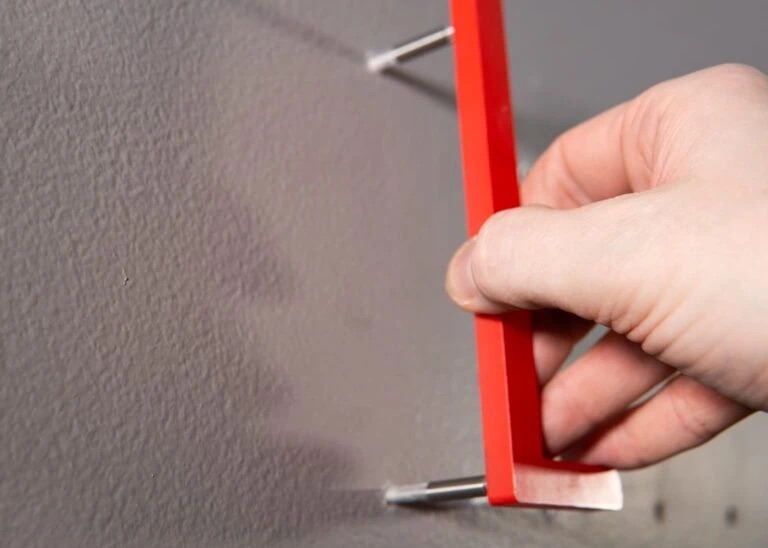 Hand hält roten Abstandshalter an Wand, um zwei Dübel/Schrauben in gleicher Höhe auszurichten.