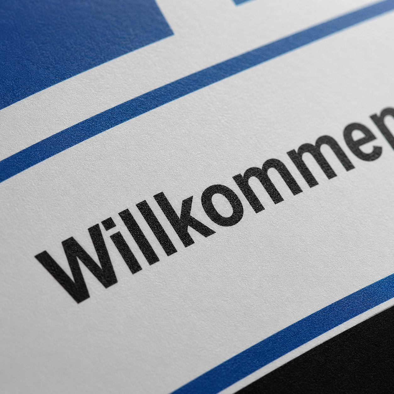 Schräg fotografierter Papierbogen mit dem Wort „Willkommen“ in schwarzer Schrift und blauen Linien als Designelemente