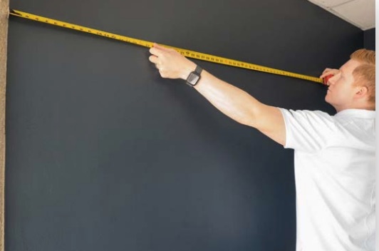 Mann in weißem T‑Shirt misst mit gelbem Maßband die Breite einer dunkelgrauen Wand bis zur Ecke.