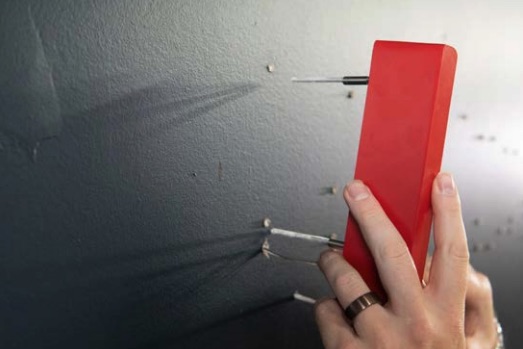 Hand hält roten Magneten an Wand, der mehrere Metallnägel anzieht und kleine Einschlagstellen zeigt.