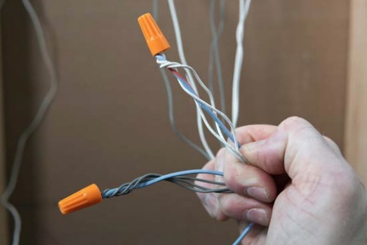 Hand hält mehrere miteinander verdrehte elektrische Leitungsdrähte mit orangefarbenen Drahtklemmen in einer Baustellenumgebung