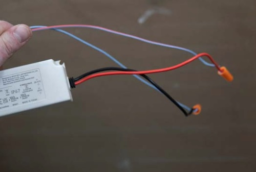 Hand hält LED-Treiber mit roten, schwarzen, blauen und rosa Kabeln, an deren Enden orange Kabelverbinder sitzen.