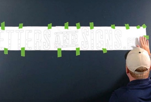 Mann mit Baseballcap klebt eine lange Papiervorlage mit Buchstaben an eine dunkle Wand, grüne Malerkreppstreifen halten sie fest.