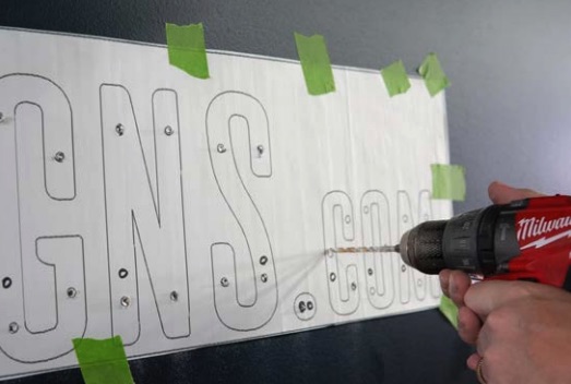 Hand hält Akkuschrauber und bohrt Löcher in rechteckiges Metallblech mit aufgemalten Buchstaben, Blech an Wand mit grünem Klebeband fixiert.
