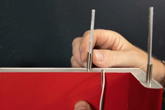 Hand hält Metallstift, der in Schraubhülse eingesetzt wird, rotes Paneel mit sichtbarem Kabel im Vordergrund.