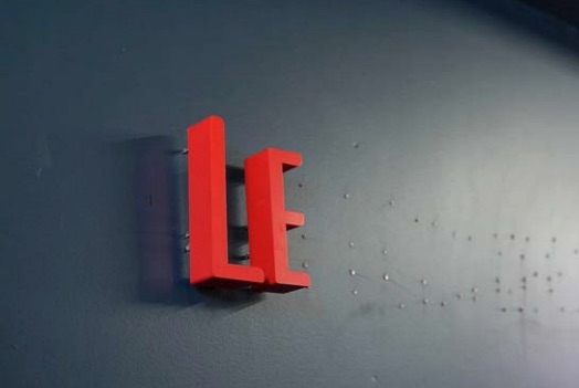 Rote 3D-Buchstaben "LE" an einer dunkelgrauen Wand, rechts Reihung kleiner Befestigungsstellen sichtbar.