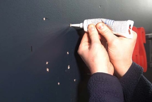 Hände drücken Dichtmittel aus einer Tube in mehrere kleine Löcher in einer dunkelblauen Wand, vermutlich zum Ausbessern.