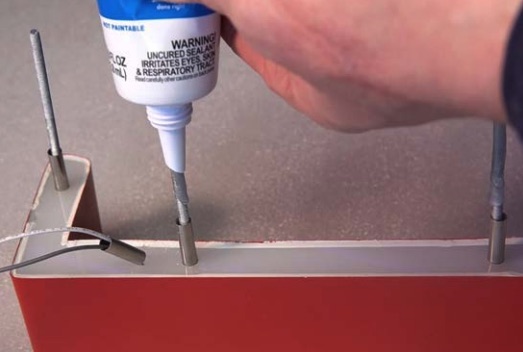 Hand spritzt Dichtungsmasse in Metallbolzen an rotem Möbel- oder Schildrahmen, sichtbare Schrauben und Kabeldurchführung.