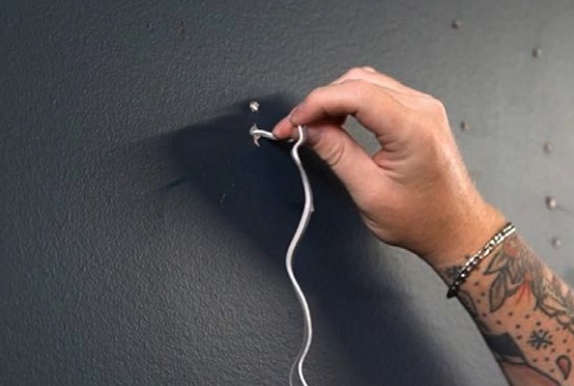 Hand mit Tätowierung und Armband hängt ein weißes Kabel an einen in Dunkelgrau gestrichenen Wandhaken.