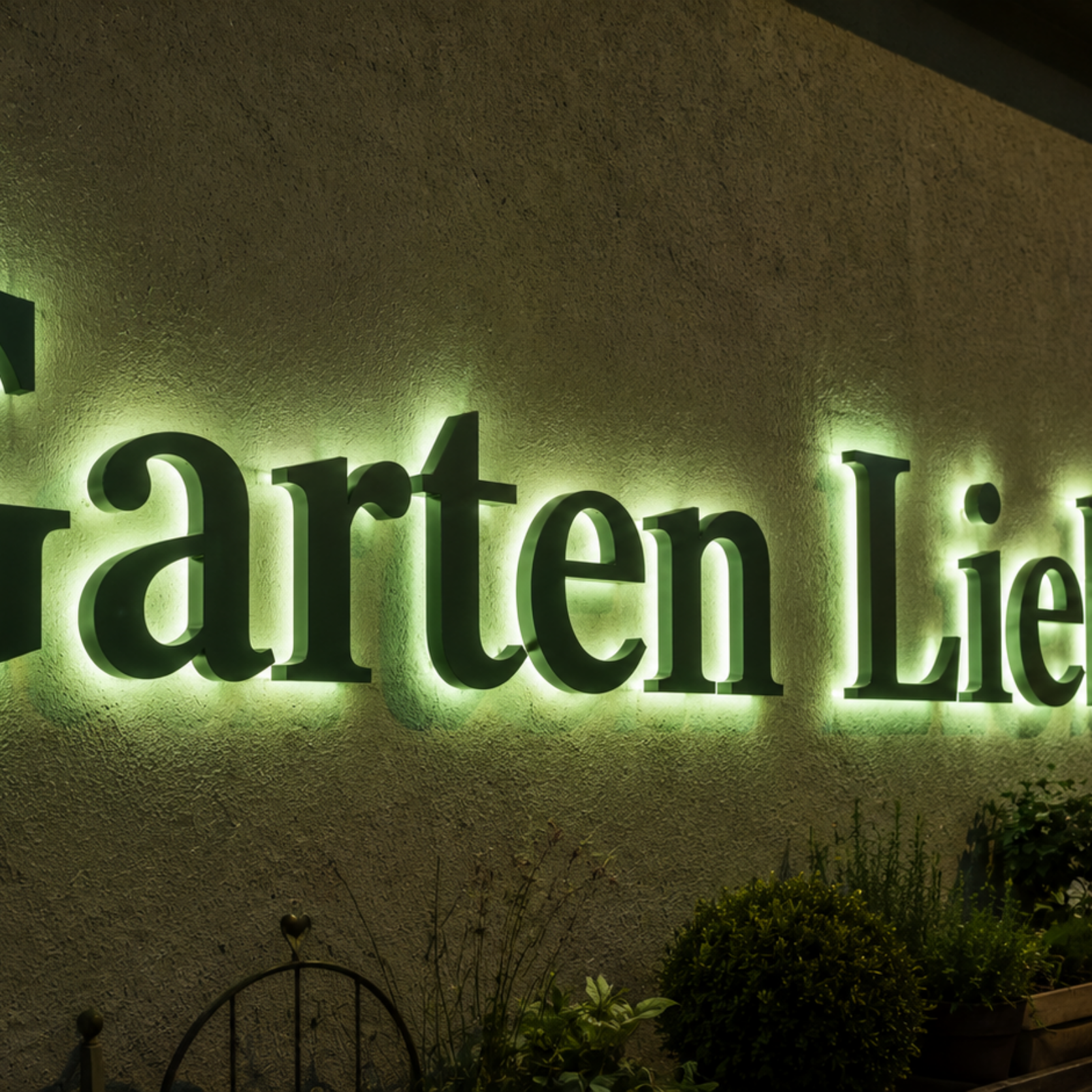 Leuchtende grüne Außenbeschriftung "Garten Liebe" an einer Hauswand über Pflanzkästen bei Dämmerung.
