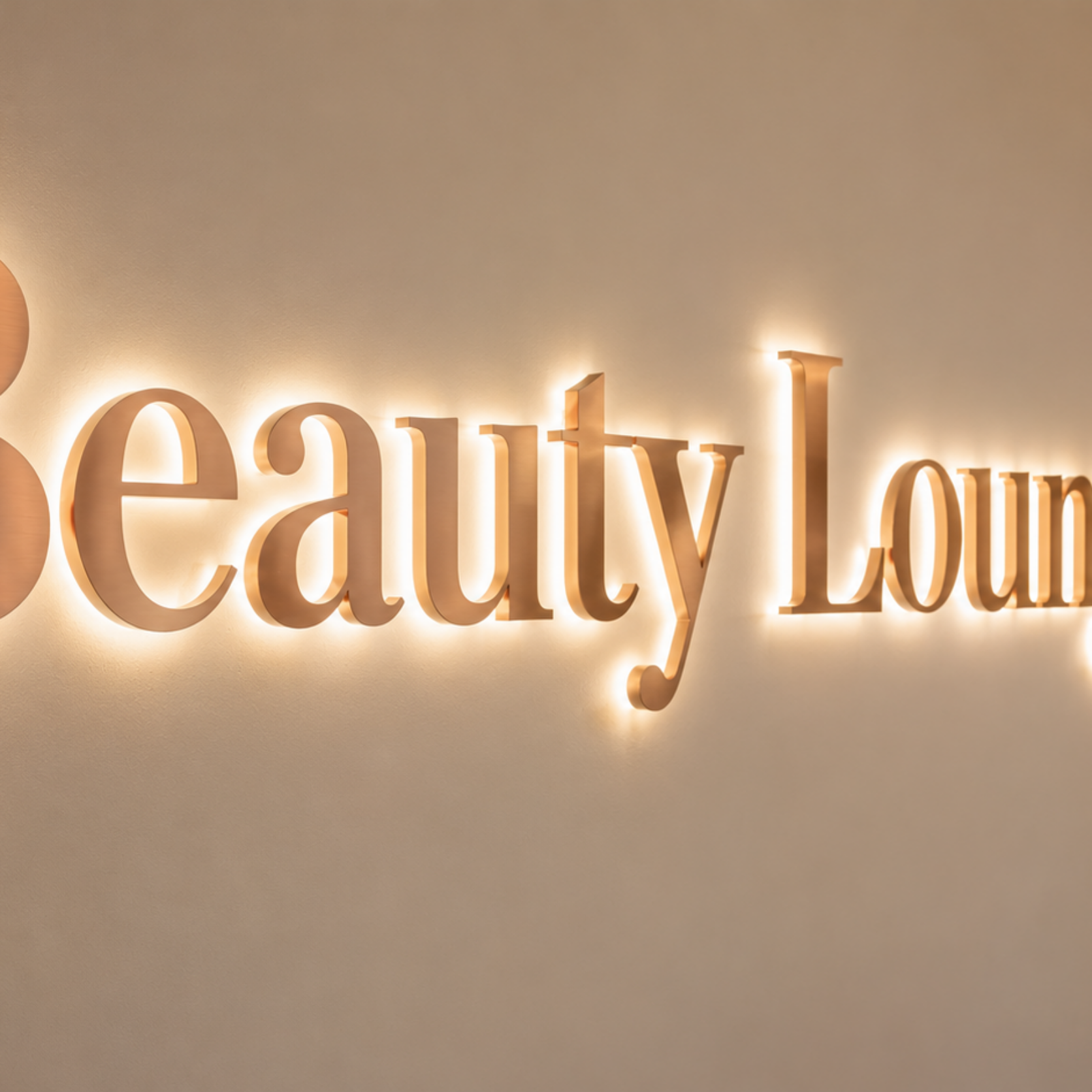 Leuchtendes Messing-Schild mit der Aufschrift "Beauty Lounge" an cremefarbener Wand, rechts Vase mit rosa Blumen auf Marmorplatte.