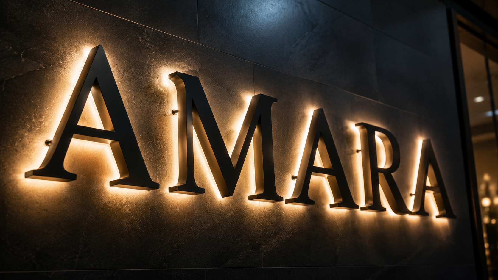 Großformatige, beleuchtete Metallbuchstaben an einer dunklen Wand, die das Wort "AMARA" mit warmem Hinterlicht zeigen.
