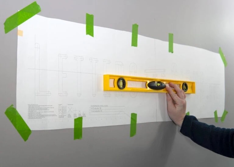 Großes weißes Papier mit technischen Zeichnungen, an einer grauen Wand mit grünem Malerband befestigt, Hand hält gelbes Wasserwaagegerät.