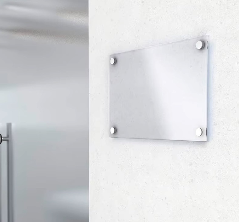 Mattes Glasnamensschild mit vier Edelstahlbefestigungen an weißer Wand, moderner Eingangsbereich im Hintergrund.