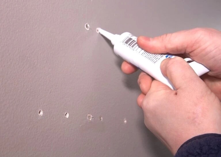Hände, die mit einer Kartuschenklebepistole Spachtelmasse in kleine Löcher an einer grau gestrichenen Wand drücken.