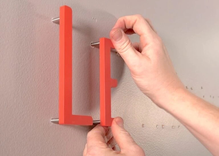 Hände montieren zwei rote, L-förmige Wandhalter mit Metallstiften an einer hellgrauen Wand.
