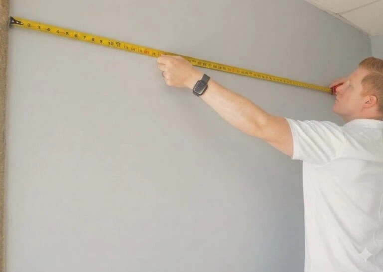 Mann in weißem T‑Shirt misst mit gelbem Maßband die Breite einer grauen Wand, Maßbandspanne sichtbar.